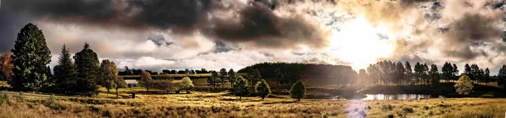 A panoramic view of a serene countryside landscape with trees, a pond, and sunlight breaking through clouds.