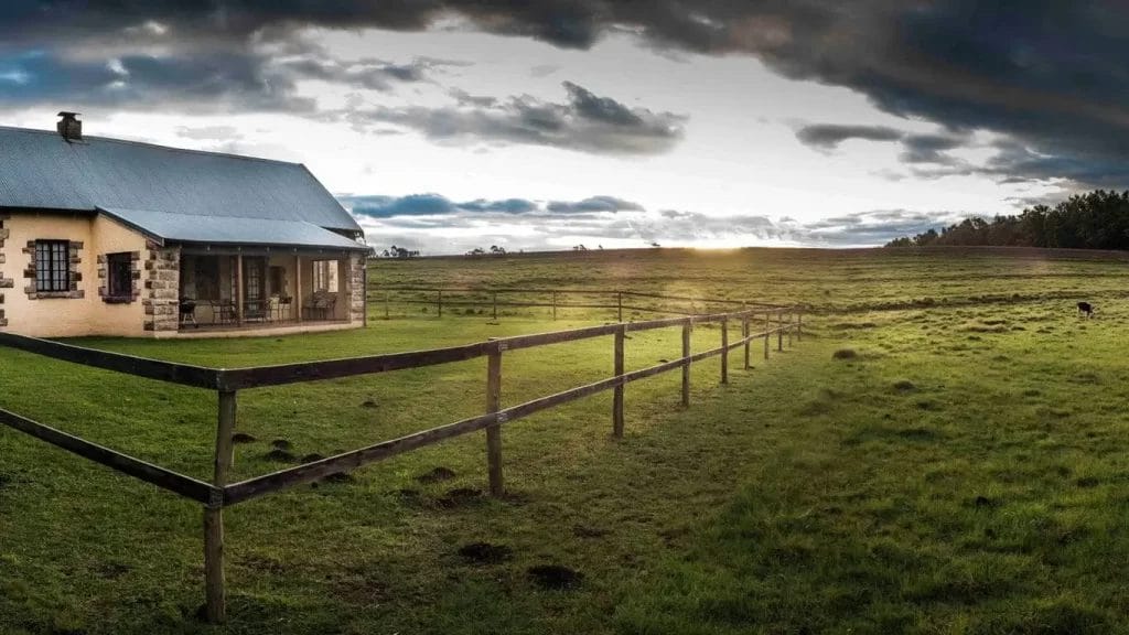 Rural landscape with a farmhouse, fenced pasture, and a setting sun piercing through clouds.