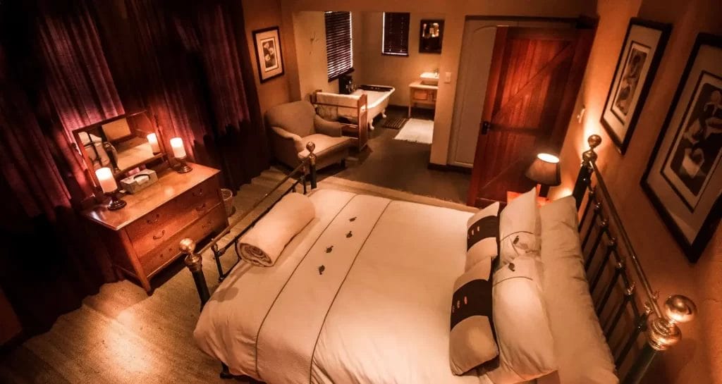 A warmly lit, cozy bedroom with an en-suite bathroom, furnished with a large bed, bedside tables with lamps, a dresser, and artwork on the walls.