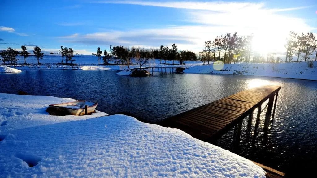 A serene winter scene with a wooden pier leading to a calm lake, surrounded by snow-covered ground and trees, under a clear blue sky.