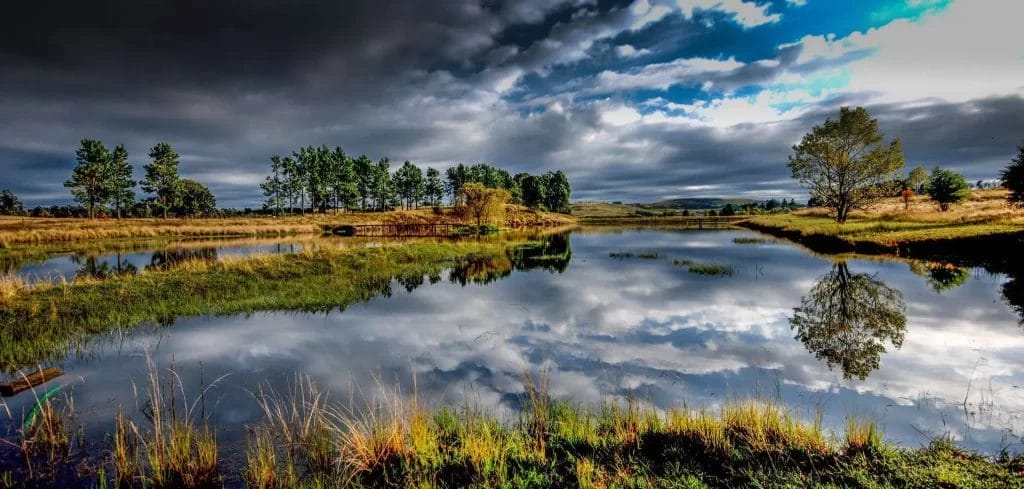 A tranquil rural landscape with a reflective pond under a dynamic sky at dusk.