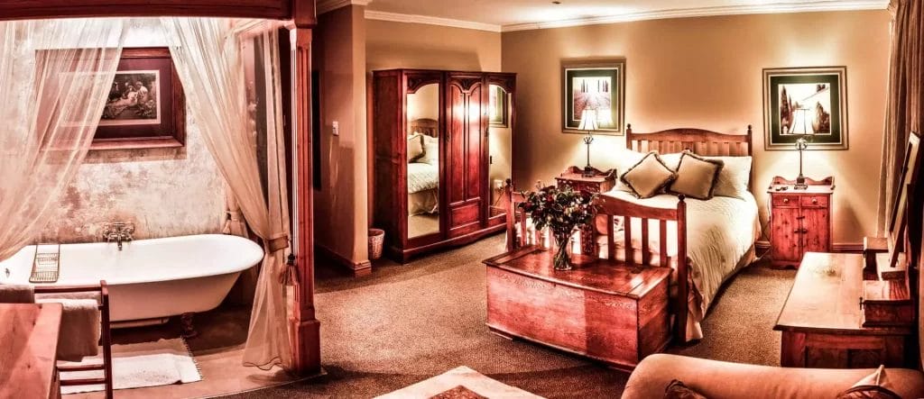 Panoramic view of an elegant and warmly lit bedroom suite with a canopy bed, freestanding bathtub, and classic wooden furniture.