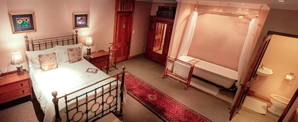 A warmly lit vintage-style bedroom with an iron bed frame, two bedside tables, and an open bathroom with a claw-foot tub.