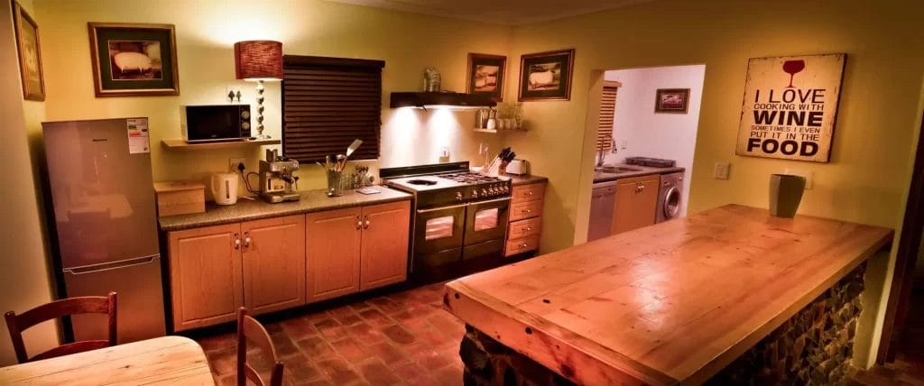 A cozy, warmly lit kitchen with wooden cabinetry and a large dining table, featuring a decorative sign about cooking with wine.