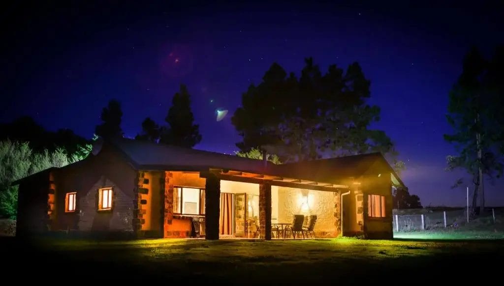 A rural house at night illuminated by interior lights with a starry sky above.
